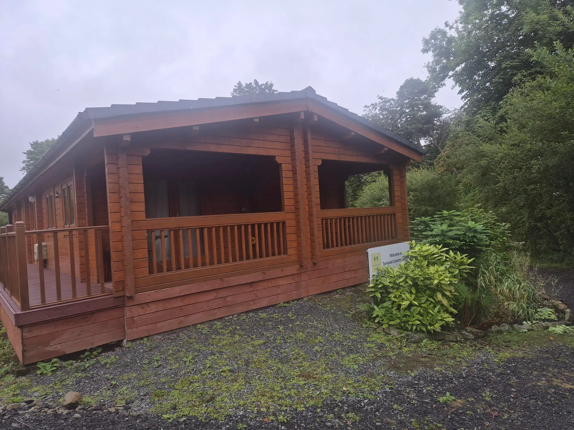 Solid timber lodge with covered veranda and welcome sign at Torwood Country Lodges, Newton Stewart